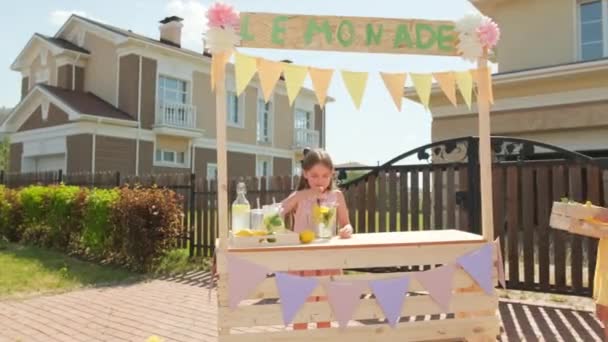 Medium shot van twee mooie 10-jarige meisjes die zich klaarmaken voor de openlucht zomermarkt staande op diy limonade stand, een roeren limonade in glas decanter en andere brengen doos citroenen - Video