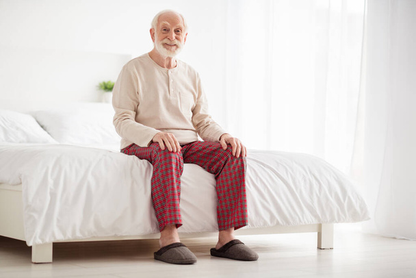 Full body photo of senior man happy positive smile sit bed home indoors wear pajamas slippers rest relax - Фото, изображение