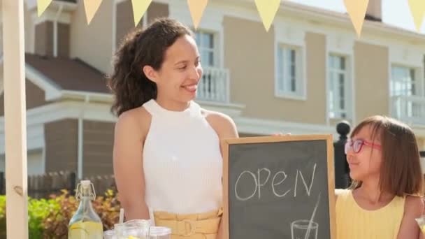 Ruční střední portrét usmívající se atraktivní smíšené rasy žena se dvěma krásnými elementární věku dcery při pohledu na kameru stojící na dřevěném stánku prodávající domácí limonádu v horkém letním dni - Záběry, video