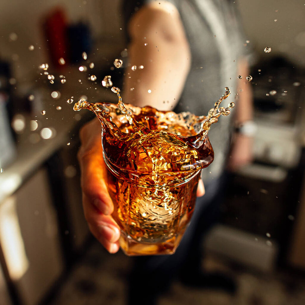 Ένας άντρας που κρατάει ένα ποτήρι ουίσκι με πιτσιλιές στην κουζίνα. - Φωτογραφία, εικόνα