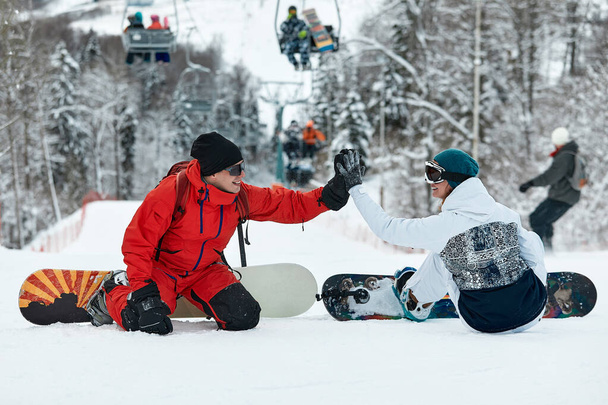 Paar Snowboarder gibt einander eine hohe Fünf, während sie auf den Schneegipfeln einer Piste vor blauem Himmel und Skilift im Hintergrund im Winterskigebiet sitzen - Foto, Bild