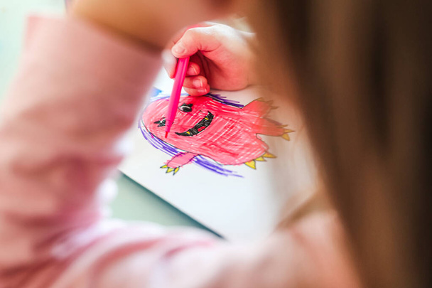 Mädchen zeichnet ein lächelndes rosafarbenes Monster mit lila Haaren, schaut unter dem Arm hervor - Foto, Bild