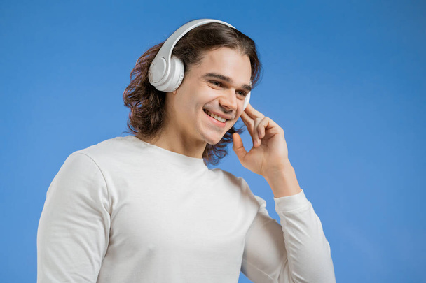 Charming young man with long hairdo listening to music with wireless headphones, guy having fun, smiling in studio on blue background. Dance, radio concept. - Foto, Bild