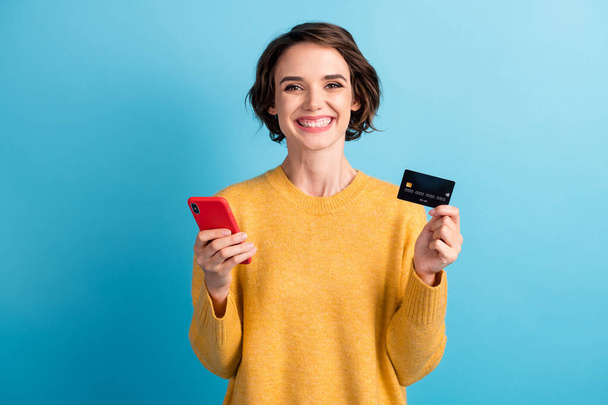 Portrait of cheerful pretty brunette bob hair young woman hold smartphone bank card dress yellow sweater isolated on blue color background - Fotó, kép