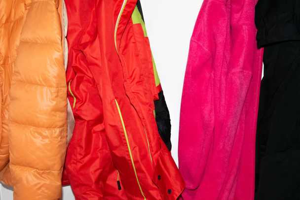 Deportes brillantes chaquetas de nylon naranja y rojo. Suéteres suaves de lana de color rosa cálido y negro, colgados en la pared blanca - Foto, imagen