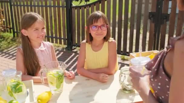 Μέτριο πλάνο δύο χαρούμενα 10χρονα κορίτσια που πουλούσαν λεμονάδα στο ύπαιθρο την ζεστή καλοκαιρινή μέρα, δίνοντας "κόλλα πέντε" το ένα στο άλλο μετά την γυναίκα που αγόραζε λεμονάδα από το περίπτερό τους - Πλάνα, βίντεο