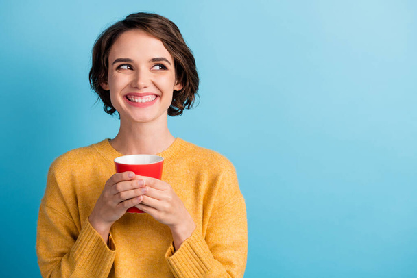 Photo of toothy beaming lady wearing casual yellow sweater holding cup hands arms looking empty space isolated blue color background - Fotó, kép