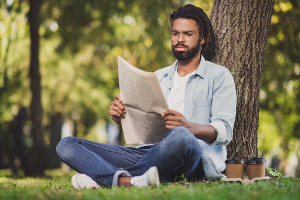 Full size photo of serious afro american man sit grass park relax read newspaper article in outdoors outside - Фото, изображение