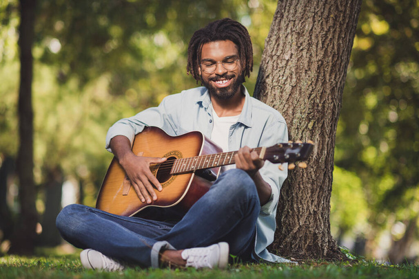 Full body photo of charming happy young dark skin man enjoy weekend sit grass play guitar outside in park outdoors - Zdjęcie, obraz