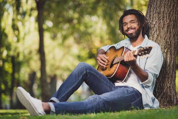 Portrait of attractive cheerful guy sitting on green grass playing guitar singing romantic hit sunny day outside outdoor - Φωτογραφία, εικόνα