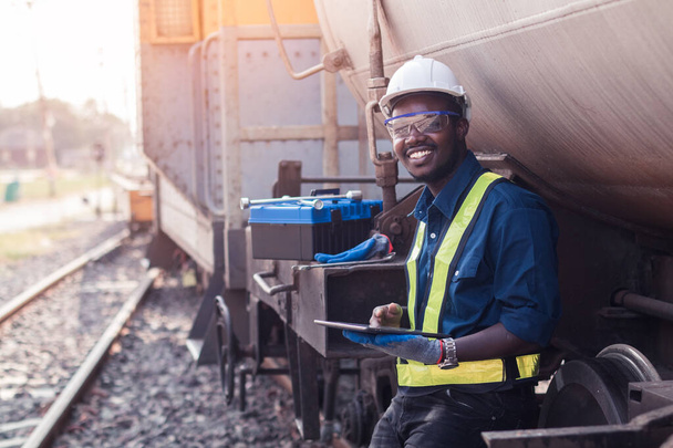 Αφρικανός τεχνικός μηχανικού φορώντας κράνος, άλση και γιλέκο ασφαλείας χρησιμοποιεί ένα κλειδί για την επισκευή του τρένου με τη χρήση tablet - Φωτογραφία, εικόνα