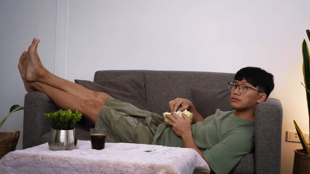Portrait of asian man in casual watching interesting movie on TV and eating popcorn while sitting on the couch in living room at home, people and entertainment concept - Foto, Imagen