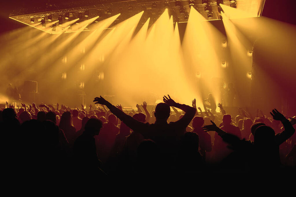 Pessoas com mãos levantadas, silhuetas de multidão de concertos na frente de luzes de palco brilhantes. - Foto, Imagem