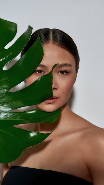 Portrait of a young magnificent half-naked asian girl in a black top face half-hidden with a monstera leaf  - Foto, afbeelding