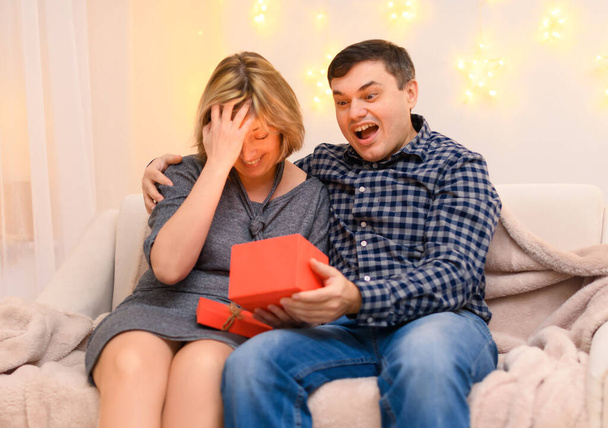 Retrato de pareja romántica adulta sentada en un sofá en casa, luces navideñas en una pared - Foto, Imagen