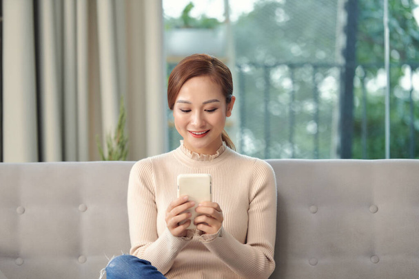 Ευτυχισμένη νεαρή Βιετναμέζα γυναίκα χαλαρώστε στο σαλόνι περιηγηθείτε ασύρματο internet στο κινητό gadget. - Φωτογραφία, εικόνα
