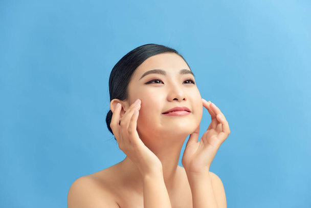 Mooie glimlachende aziatische meisje model met natuurlijke make-up aanraken gloeiende gehydrateerde huid op blauwe achtergrond closeup - Foto, afbeelding