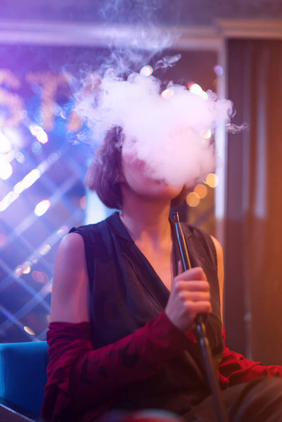Une femme méconnaissable dans une boîte de nuit ou un bar fume un narguilé ou une chicha. Le plaisir de fumer. Fumée sexy avec vape - Photo, image