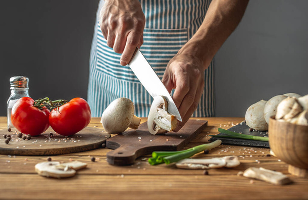 A male chef in a blue apron is cutting fresh mushrooms with a knife. Concept of process of cooking a delicious dish - Photo, Image