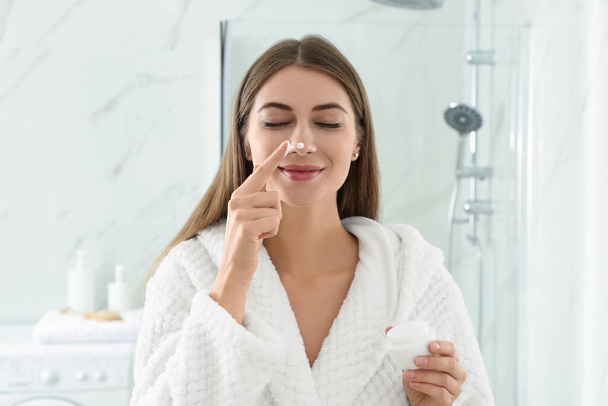 Young woman applying face cream onto her nose in bathroom - Φωτογραφία, εικόνα