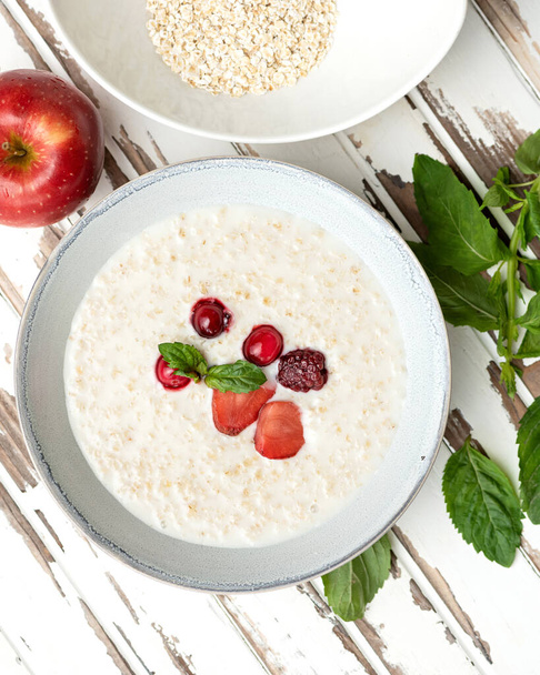 Oatmeal with milk and berries. Nutritious breakfast, wholesome healthy food. Bowl of porridge cereal oats with fresh red berries and mint on on white wooden rustic table. High angle view - Photo, Image