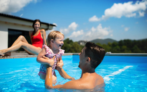 Młoda rodzina z małą córeczką w basenie na świeżym powietrzu w ogrodzie podwórkowym, zabawy. - Zdjęcie, obraz