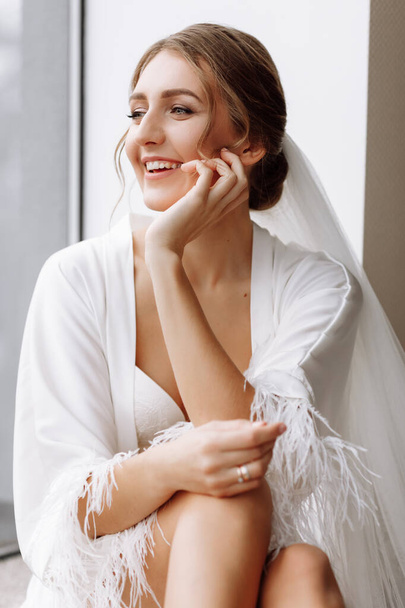 花嫁の朝。豪華な花嫁だ。窓の白いローブに長い髪の美しい女性。結婚式の前の花嫁の朝。自然の美しさとプロの化粧。スキンケア製品 - 写真・画像