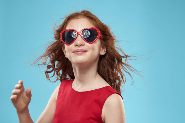 Little girl in sunglasses in the form of hearts red dress curly hair blue background - Photo, Image