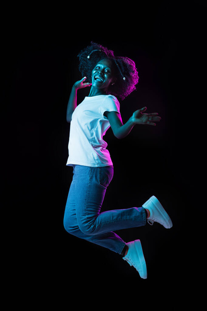 Genç Afro-Amerikalı kız, neon ışıklı karanlık arka planda izole bir şekilde atlıyor.. - Fotoğraf, Görsel