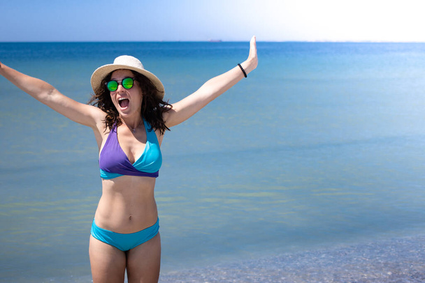 Felice eccitato giovane donna a braccia aperte in piedi sulla spiaggia sul mare in costume da bagno bikini blu e cappello estivo, bocca aperta in amazement.Godersi la vita in paradiso.Stagione di vacanza 2021.Copia lo spazio per la compagnia di viaggi  - Foto, immagini