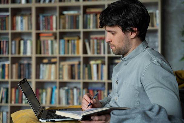Portrait of a man sitting on a sofa with a laptop on the background of a bookshelf - Φωτογραφία, εικόνα