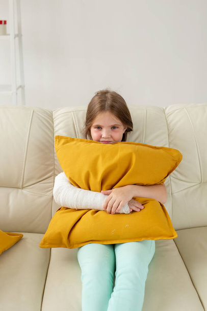 Dziecko z gipsem na złamanym nadgarstku lub ramieniem siedzącym na kanapie. Koncepcja odzyskiwania i choroby. - Zdjęcie, obraz