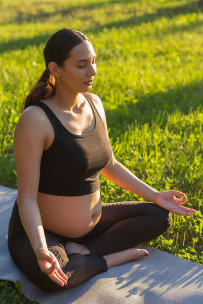Cute young caucasian pregnant woman is meditating while sitting on a rug on the lawn on a sunny summer evening. Concept of pacification and energy boost - Foto, afbeelding