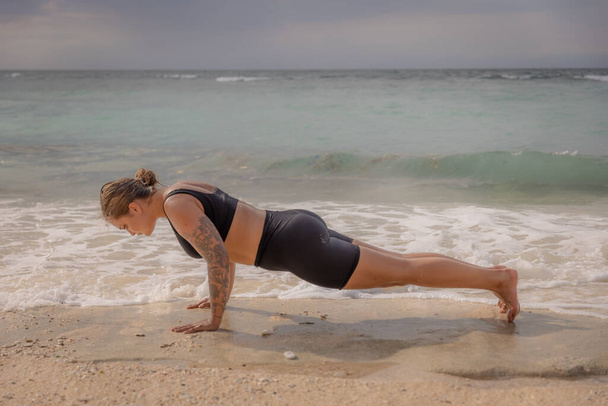 ビーチでヨガの女性。白人女性の練習Phalakasana,プランクポーズ.強い体だ。健康的なライフスタイル。セルフケアの概念。ヨガの隠れ家だ。インドネシアのバリのトーマスビーチ - 写真・画像