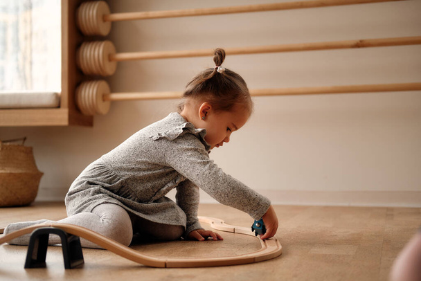 Little girl in a gray dress plays in the childrens room with a wooden railroad and a train. He is holding a wooden train with a blue and green waganomi. Educational wooden toys for children.  - Valokuva, kuva