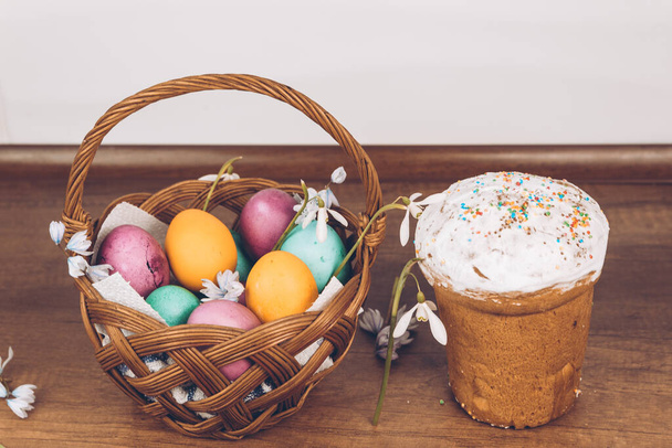 Fond de Pâques. Œufs de Pâques dans un panier et pâtisseries de Pâques. Kulich. Sur une table en bois se trouve un panier de Pâques avec des œufs jaunes, verts et violets, décoré de gouttes de neige. - Photo, image