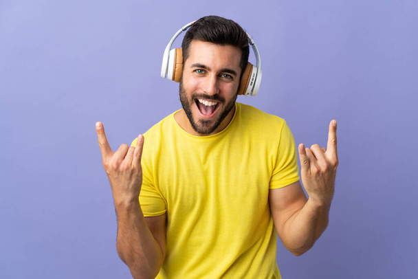 Joven hombre guapo con barba aislada sobre fondo púrpura escuchando música haciendo un gesto de rock - Foto, Imagen