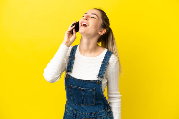Młoda kobieta korzystająca z telefonu komórkowego na odizolowanym żółtym tle śmiejąc - Zdjęcie, obraz