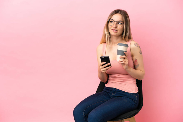 Młoda kobieta siedzi na krześle na odizolowanym różowym tle trzymając kawę do zabrania i komórkę myśląc coś - Zdjęcie, obraz
