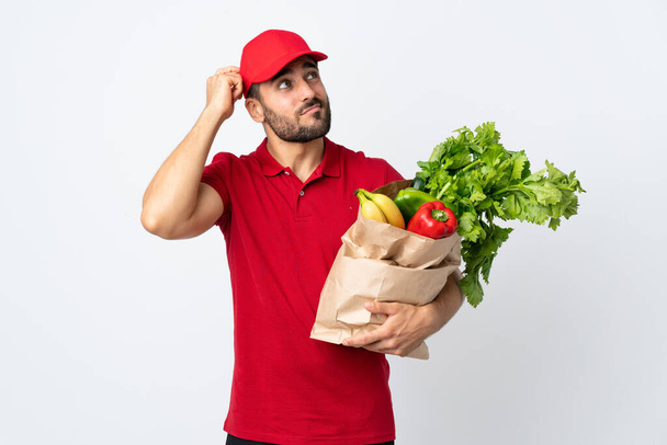 Young man with beard holding a bag full of vegetables isolated on white background having doubts and with confuse face expression - Photo, Image