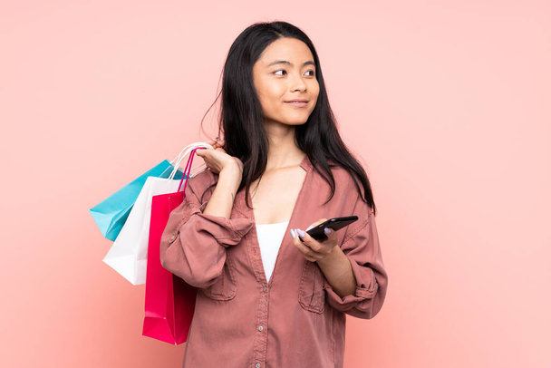 ティーネージャー中国の女の子絶縁オンピンクの背景保持ショッピングバッグと携帯電話 - 写真・画像
