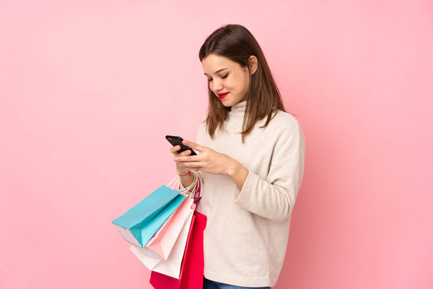 Девушка-подросток, изолированная на розовом фоне, держит сумки с покупками и пишет сообщение на мобильный телефон другу - Фото, изображение
