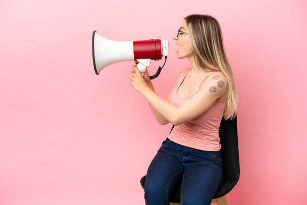 Young woman sitting on a chair over isolated pink background shouting through a megaphone - Fotografie, Obrázek
