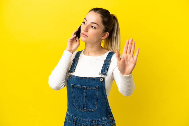 fiatal nő mobiltelefon felett elszigetelt sárga háttér így stop gesztus és csalódott - Fotó, kép