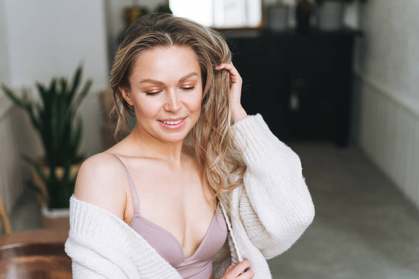 Blonde smiling woman 35 year clean fresh face and hands with long hair in lingerie and cozy knitted cardigan at the bright interior - Фото, изображение