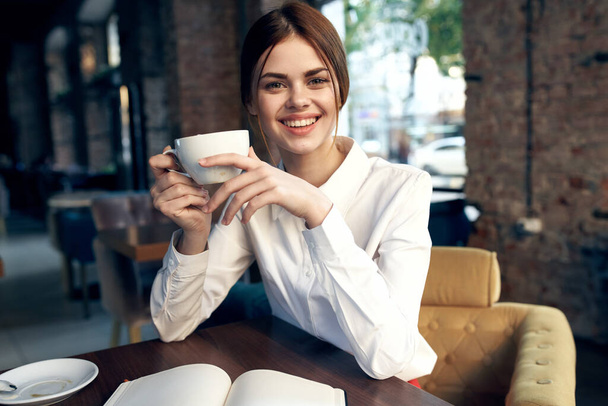 mujer feliz con una taza en la mano se sienta en una silla en el restaurante y el interior en el fondo - Foto, imagen