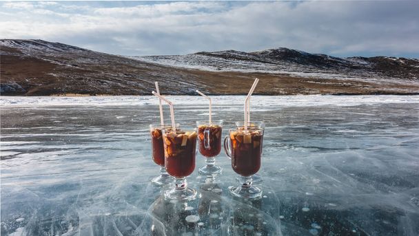 Glass mugs with mulled wine stand on a frozen lake. Red wine, sliced fruits, tubules. The ice is transparent, with cracks and gas bubbles in the depths. Background - snow-covered hills, sky. Baikal - Photo, Image