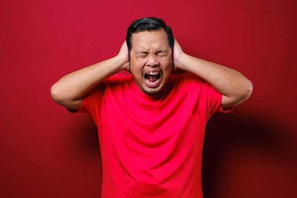 Portrét mladého asijského muže v ležérní červené košili zavírající oči a uši nechtějí nebo vyhnout se slyšet špatné zprávy, stres pod mentálním tlakem koncepce nad červeným pozadím - Fotografie, Obrázek