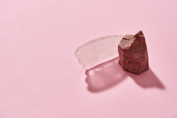Close up of natural raw pink rose quartz rough stone and Gua Sha scraper for facial massage isolated over pink background - Photo, Image