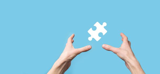 Male hand holding puzzle icon on blue background. pieces representing the merging of two companies or joint venture, partnership, Mergers and acquisition concept - Photo, Image